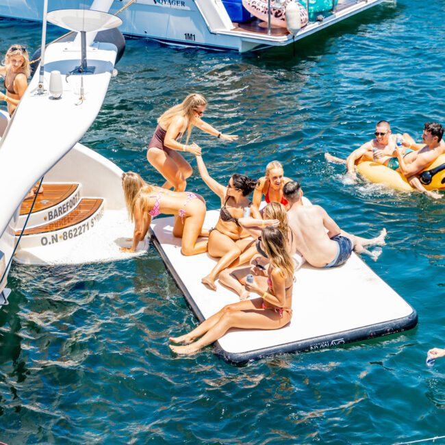 A group of people enjoying a sunny day on a floating platform in the water, surrounded by boats. Some are sitting, while others are lounging on inflatables. The atmosphere is lively and festive.