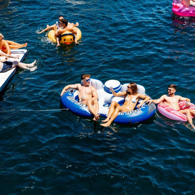 People relaxing on inflatable floats in a body of water. Two lie on a blue "Fiji" float, another person on a yellow duck, and others on pink floats. Boats are nearby, and the scene is lively and sunny, with clear blue water.