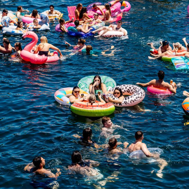 A vibrant scene of people enjoying a sunny day on the water with colorful inflatable pool floats. The clear blue water hosts various themed floats, including a flamingo and a donut. A boat with more people is anchored nearby.