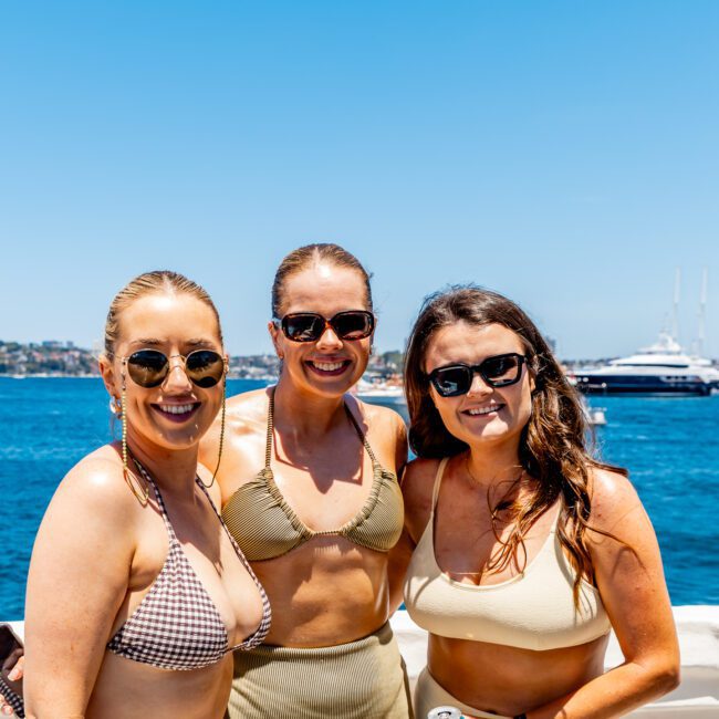 Three women in swimsuits and sunglasses smile at the camera on a sunny day by the water. Boats and a clear blue sky create a scenic background.
