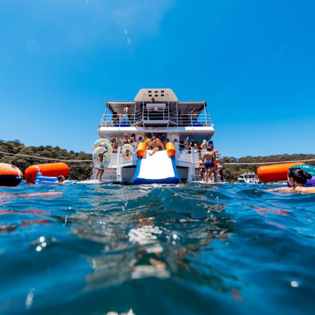 A yacht is anchored on water with people enjoying a sunny day. Some individuals are in the water near orange flotation devices. Others are on the yacht, with a slide extending into the water. Hills and clear blue skies are in the background.
