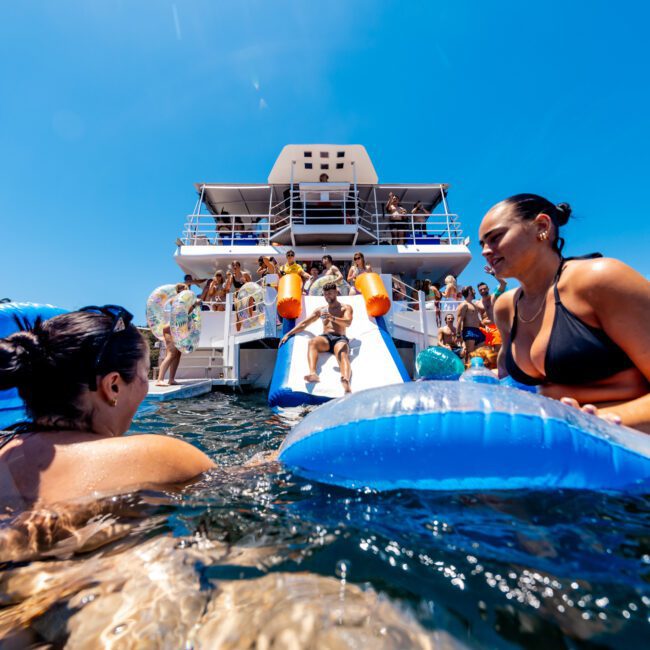 People enjoying a sunny day on a yacht with inflatable pool toys and a water slide. The water is clear, and the sky is bright blue. The yacht is crowded with people socializing and having fun.