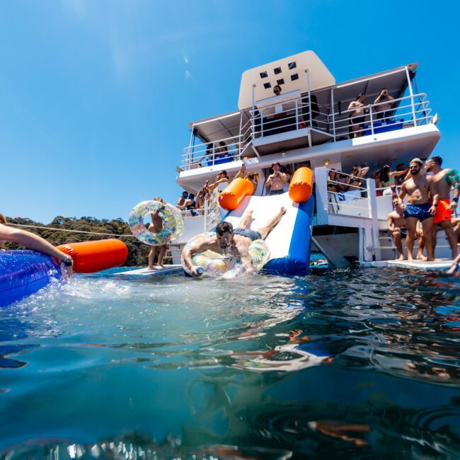 People enjoying a sunny day on the water, sliding down inflatables from a yacht into the sea. Others relax on floating devices nearby. The scene is lively, with blue skies and clear water.