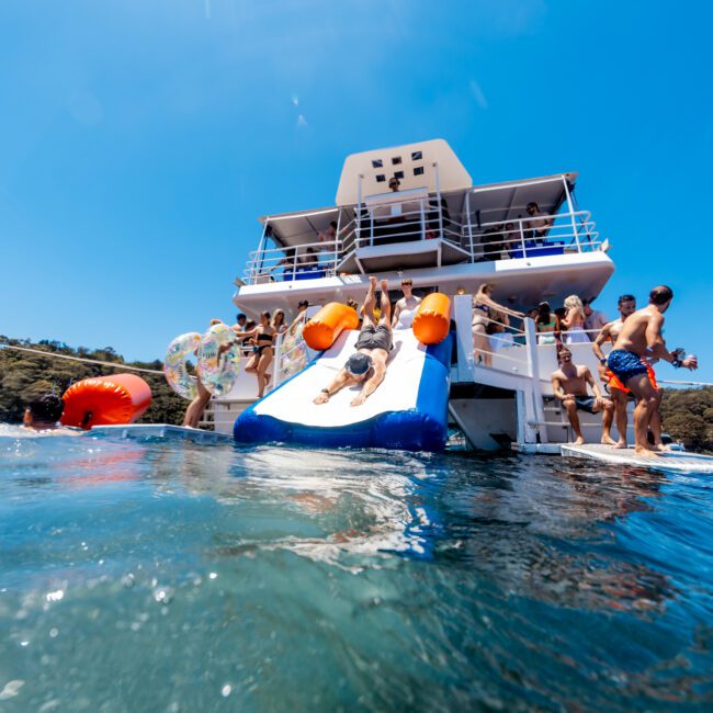 A yacht party with people enjoying the sunshine. Some are on the boat, while others slide into the water from an inflatable slide. The clear blue sky and water create a vibrant, festive atmosphere.