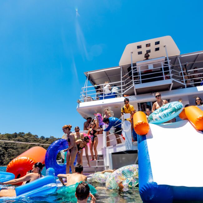 A group of people enjoy a sunny day on a boat with a slide leading into the water. Some are swimming while others relax on the boat's deck. The sky is clear and blue, enhancing the vibrant atmosphere.