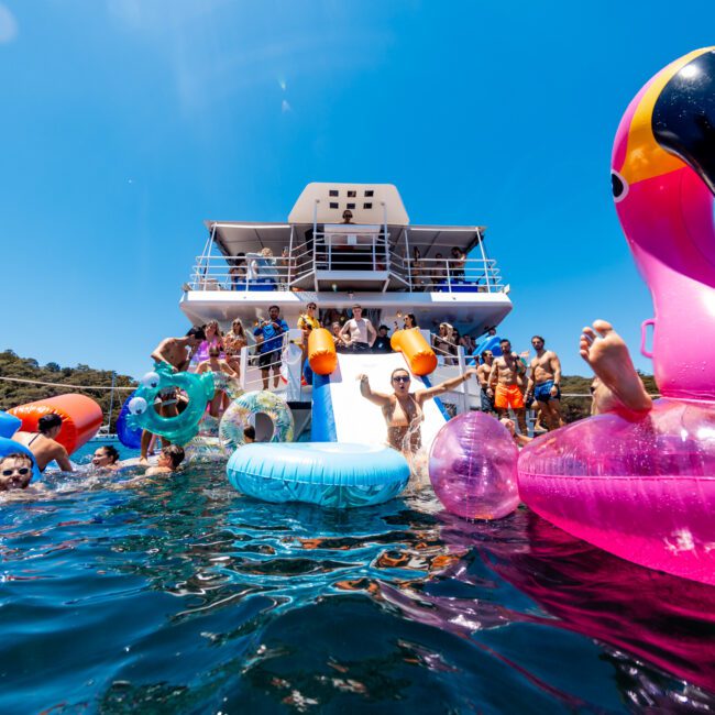 People enjoying a sunny day on a yacht, swimming and playing with colorful inflatable floats in the water, including a pink flamingo. The sky is clear and blue, and the scene is vibrant and lively.