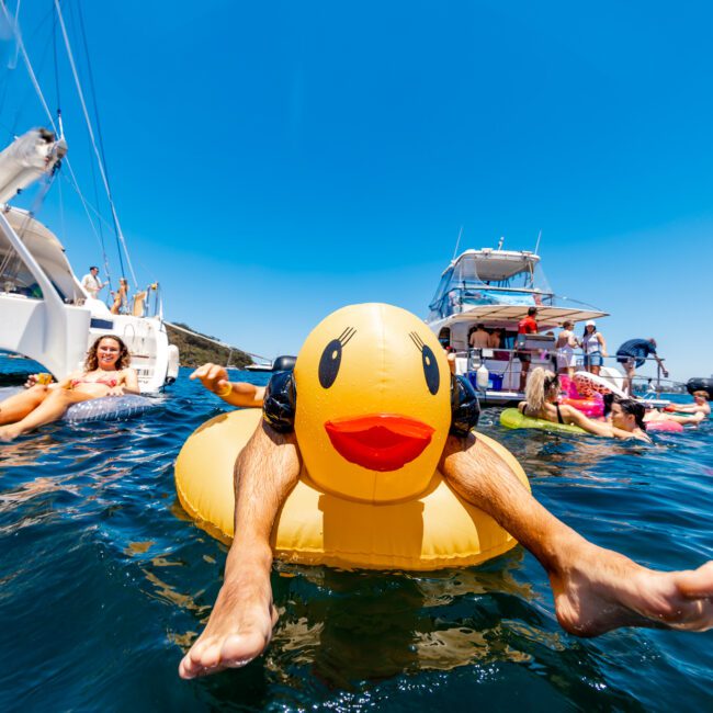 People are enjoying a sunny day on the water, surrounded by boats. A person is floating on an inflatable duck with their legs sticking out. Other attendees are relaxing and socializing on nearby boats. The words "The Yacht Social Club" are visible in the corner.