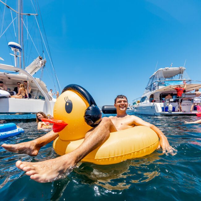 A man is floating on a large inflatable duck in the sea, surrounded by other people on floats and yachts under a clear blue sky. The scene is lively and relaxed, with a festive atmosphere.