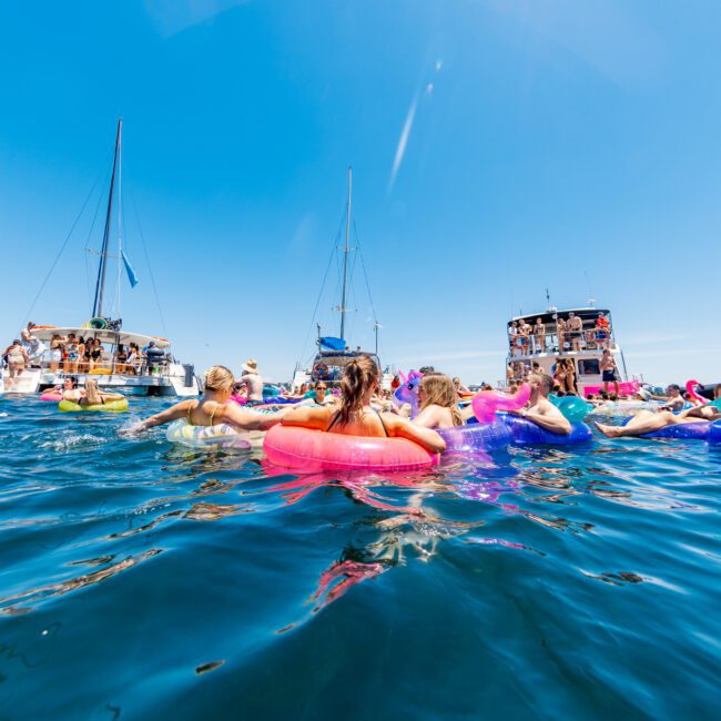 A group of people floats on colorful inflatable rings in the ocean, having fun under a clear blue sky. Several boats are anchored nearby, with more people on board enjoying the sunny day.