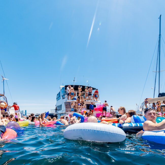 A crowded beach party scene with people floating on inflatable rings in the water near a boat. Many individuals are sunbathing and socializing under a clear blue sky. A man wears a straw hat while others enjoy the sunny day.