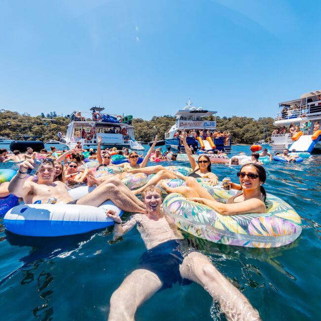 People are enjoying a sunny day on inflatable floats in a clear blue body of water. Several boats are anchored nearby with more people visible on board, creating a lively and festive atmosphere.