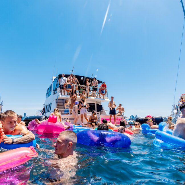 A lively scene with people enjoying a sunny day on the water. Several individuals are floating on colorful inflatable rafts near a yacht, while others are dancing or relaxing on the boat. The sky is clear and blue.