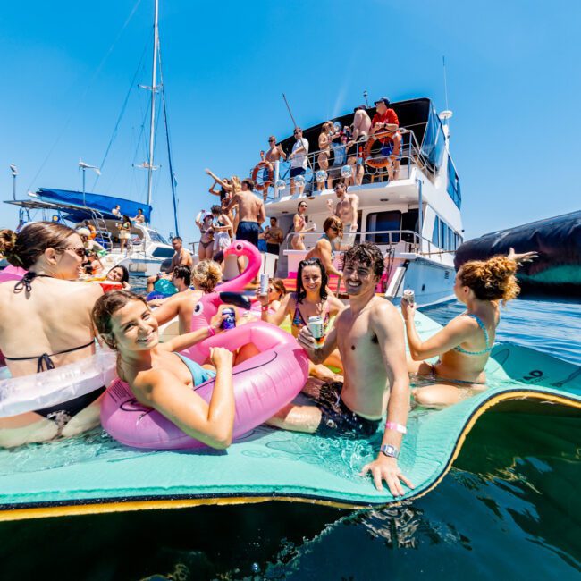 A group of young adults enjoying a sunny day on the water, floating on a large mat with inflatable toys. More people are partying on a nearby boat. The sky is clear and the atmosphere is lively and cheerful.
