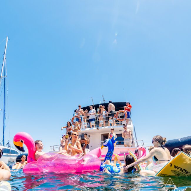 A group of people enjoying a sunny day on a yacht and in the water. Some are on inflatable toys, like a pink flamingo, while others relax on the boat. The sky is clear and blue, adding to the lively and festive atmosphere.
