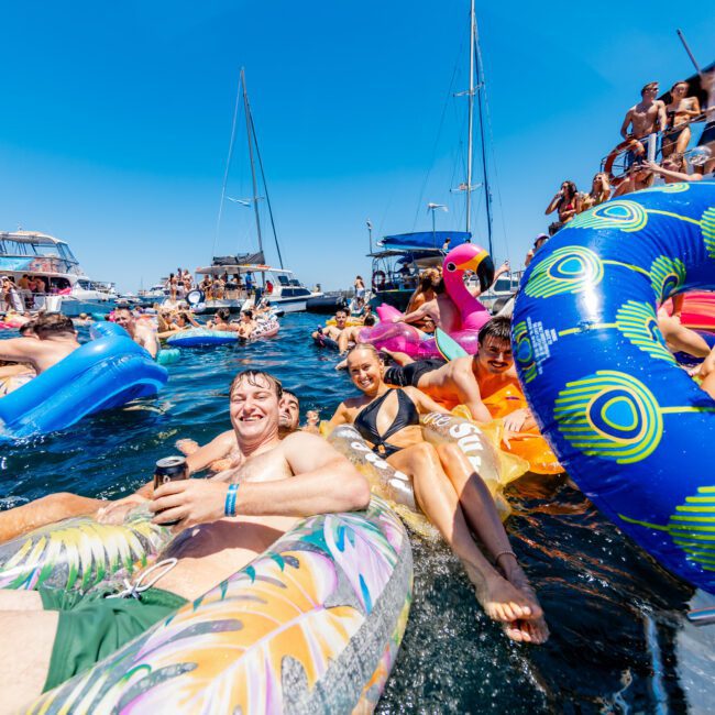 A lively scene of people relaxing on colorful inflatables in the water, surrounded by boats under a clear blue sky. Many are smiling and enjoying the sunny day at what appears to be a social yacht event.