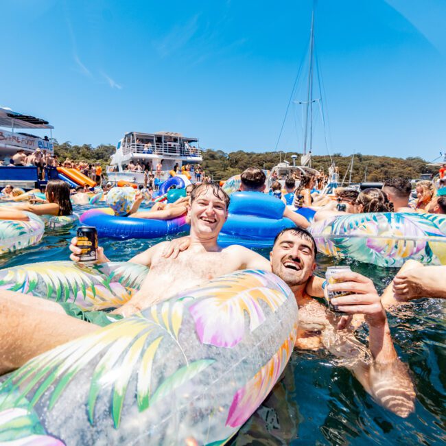 A large group of people on inflatable tubes enjoy a sunny day in the water surrounded by boats. Two men in the foreground hold drinks and smile at the camera. The scene is lively and festive, with clear blue skies above.