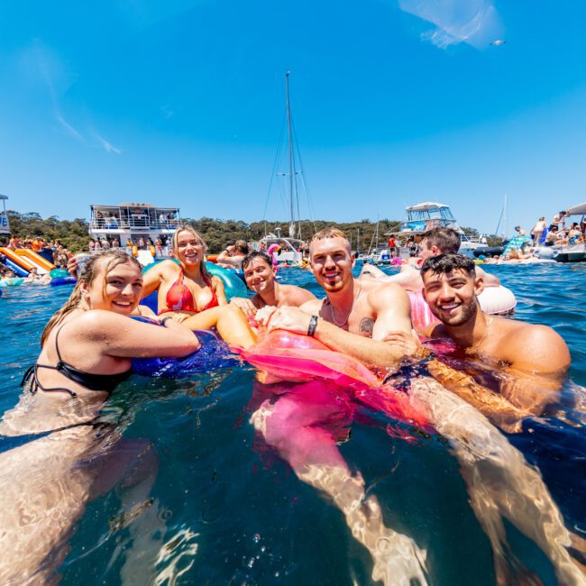 A group of smiling people in swimwear enjoy the water, holding a large inflatable float. Boats are in the background under a clear blue sky. It appears to be a lively and fun gathering on a sunny day.