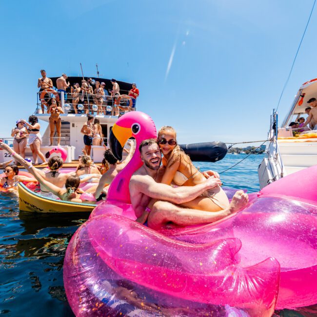 A couple sits on a pink flamingo float in the water, surrounded by others enjoying a yacht party. Two large boats are in the background, filled with people socializing and sunbathing under a clear blue sky.