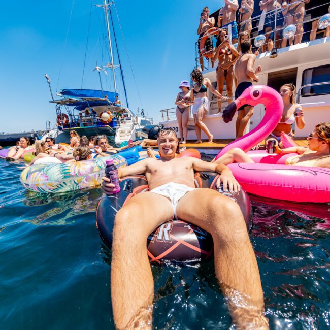 A man relaxes on a float in the water, surrounded by colorful inflatable pool floats shaped like flamingos and donuts. Other people are seen enjoying themselves on a large boat and in the water under a clear blue sky.