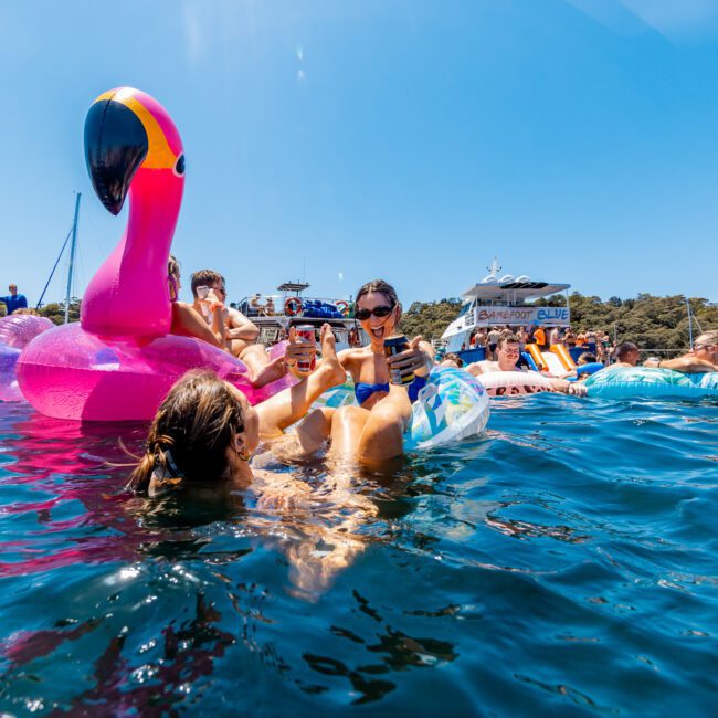 People enjoying a sunny day on the water, floating on colorful inflatables, including a large pink flamingo. They are taking photos and surrounded by boats and other inflatables in a lively, festive atmosphere.