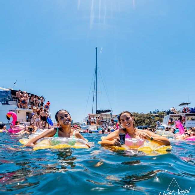 Two women are smiling while floating on inflatable rings in the water, surrounded by many people enjoying a sunny day near anchored boats. The scene is lively, with clear blue skies and the words "The Yacht Social Club" visible in the lower right corner.
