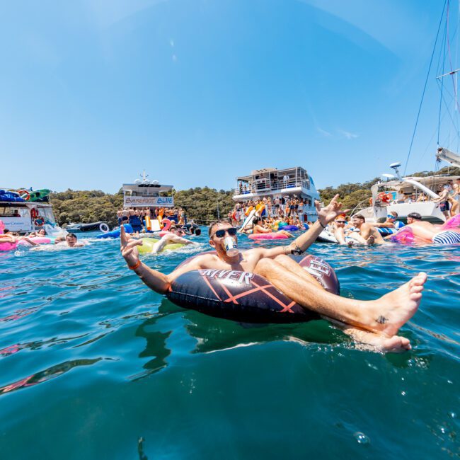 People are floating on inflatable tubes and boats in clear blue water, enjoying a sunny day. Some boats have groups of people partying. The scene is lively and festive, with lush green trees in the background.