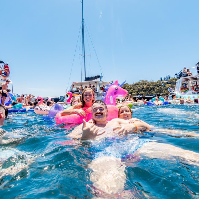 A group of people enjoying a sunny day in the ocean, floating on inflatable toys near anchored yachts. A person in the foreground holds a pink unicorn float, smiling with others around. The sky is clear and blue.