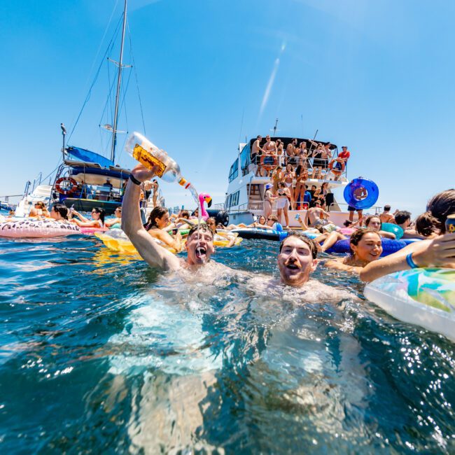 People are enjoying a lively boat party on the water under a clear blue sky. Two men in the foreground are swimming, holding drinks, and smiling. Boats and more partygoers are in the background, along with inflatables.