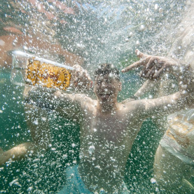 A person swimming underwater holding a bottle with playful hand gestures. Bubbles and sunlight create a vibrant, dynamic scene. Other swimmers are partially visible in the background. The water is clear and the atmosphere lively.