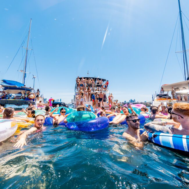 A lively scene of people enjoying a sunny day on the water, floating on colorful inflatables near boats. The sky is clear and blue. A crowd gathers on a large boat, adding to the festive atmosphere.