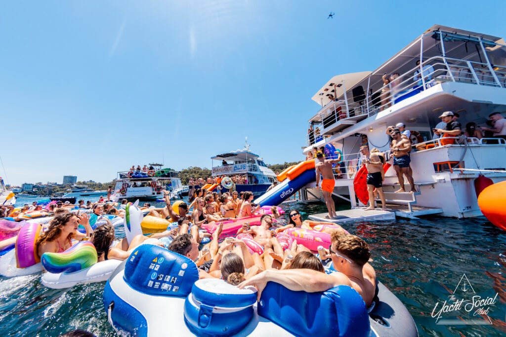 A lively boat party scene shows a large group of people enjoying the sun and water. Many are on colorful inflatable tubes and rafts, around and near a boat. The sky is clear, and other boats are visible in the background.