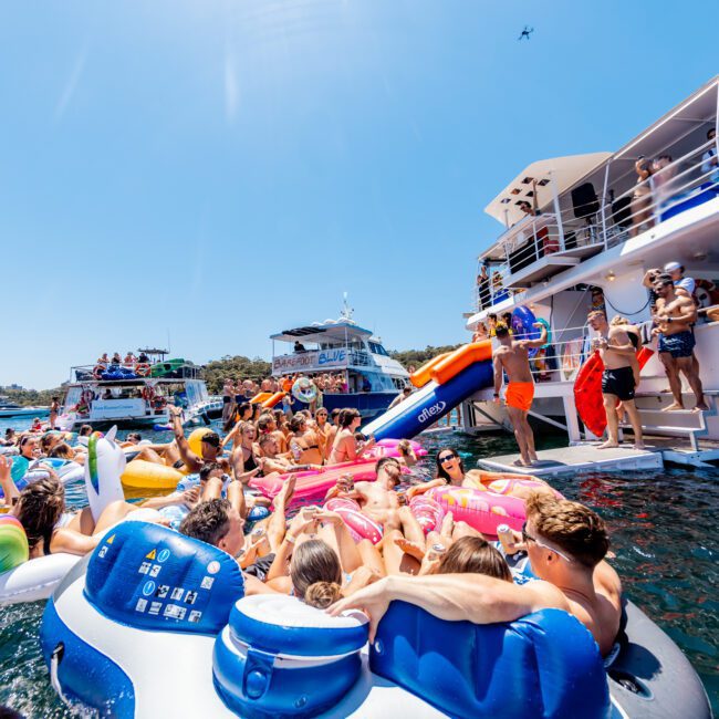 A lively boat party scene shows a large group of people enjoying the sun and water. Many are on colorful inflatable tubes and rafts, around and near a boat. The sky is clear, and other boats are visible in the background.