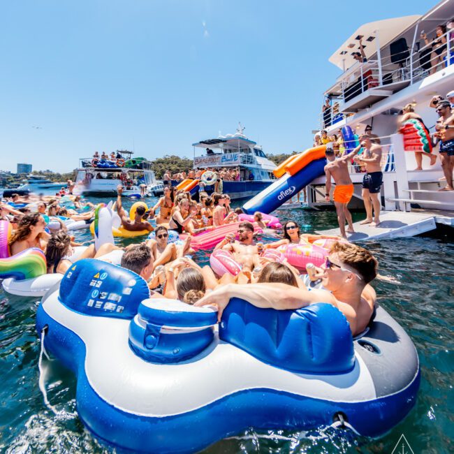 A lively scene of people enjoying a sunny day on the water, with many floating on inflatable rafts. Boats are anchored nearby, and a waterslide extends from a yacht. The atmosphere is festive and relaxed.