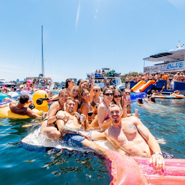A group of people in swimsuits are floating on inflatable rafts in a sunny lake. They are smiling and holding drinks. Behind them, several boats and more people on inflatables are visible, enjoying the water and clear sky.