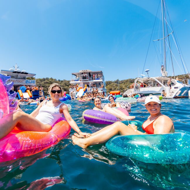A group of people relaxing on inflatable pool floats in the water near anchored boats. They are smiling, with a clear blue sky above and lush greenery in the background. Some floats are shaped like an unicorn and a doughnut.