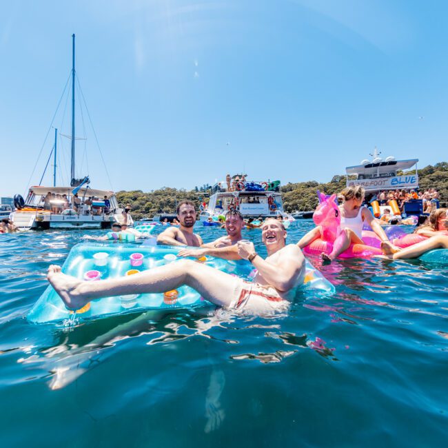 A group of people relaxing on inflatable floats in a sunny, clear blue sea with boats and yachts in the background. Pink inflatable flamingos are visible, and everyone seems to be enjoying a festive atmosphere.