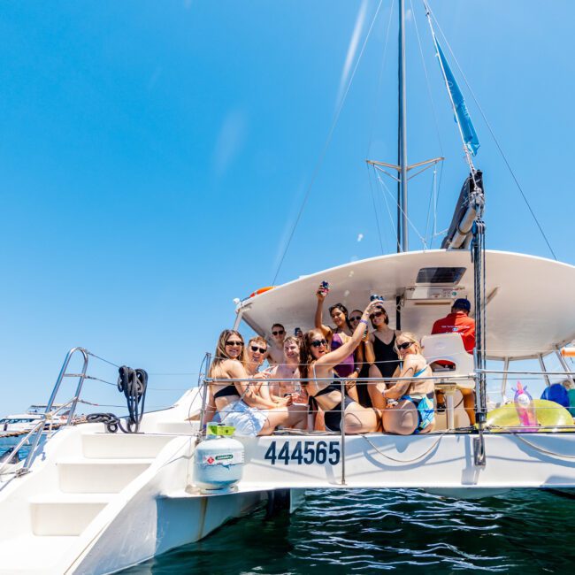 A group of people are sitting on the back deck of a catamaran yacht, enjoying the sunny day. They are smiling and taking photos. The boat is surrounded by clear blue water, and other boats are visible in the distance under the clear sky.