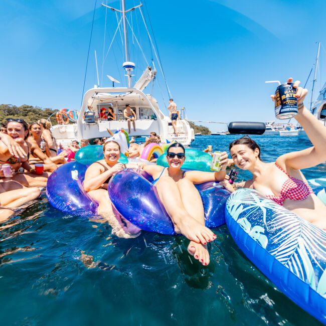 A group of people are floating on inflatable rafts and swimming in the ocean near anchored boats. They are enjoying drinks and wearing swimwear under a clear blue sky. The scene suggests a lively and fun summer gathering.