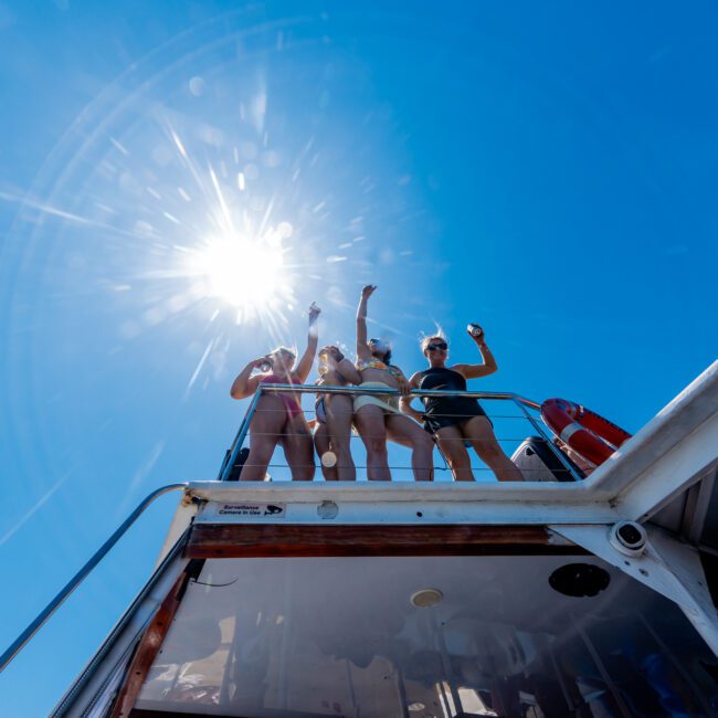 Four people in swimwear stand on the deck of a boat under a bright blue sky, celebrating with raised arms. Sunlight shines brightly, casting lens flares.