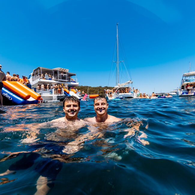Two people swimming in the ocean, smiling at the camera, surrounded by several boats with others swimming and relaxing. The sky is clear and sunny. The words "The Yacht Social Club" appear in the bottom right corner.