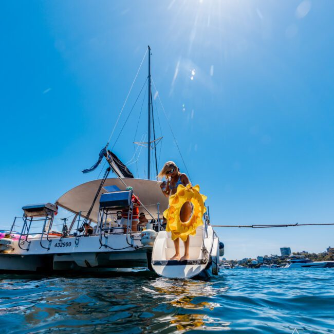 A person wearing a yellow flotation ring stands on the lower deck of a sailboat under a bright, sunny sky. The sailboat is anchored on calm blue waters with land visible in the background.
