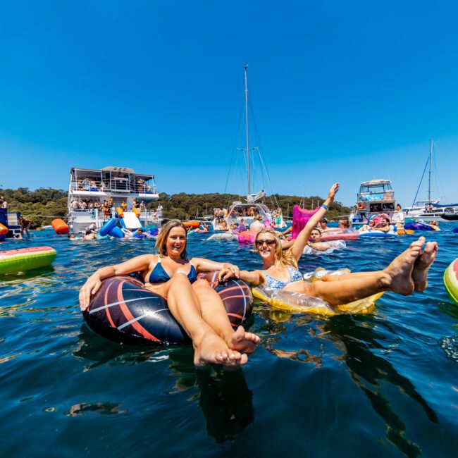 Two people smiling and lounging on inflatable floats in a lake, surrounded by others enjoying the water. Boats and jet skis are visible in the background under a clear blue sky.
