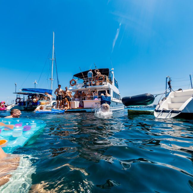 A lively scene of people enjoying a sunny day on the water. Several boats are anchored, with groups of people swimming and lounging on floaties. The sky is clear and blue, adding to the vibrant, festive atmosphere.