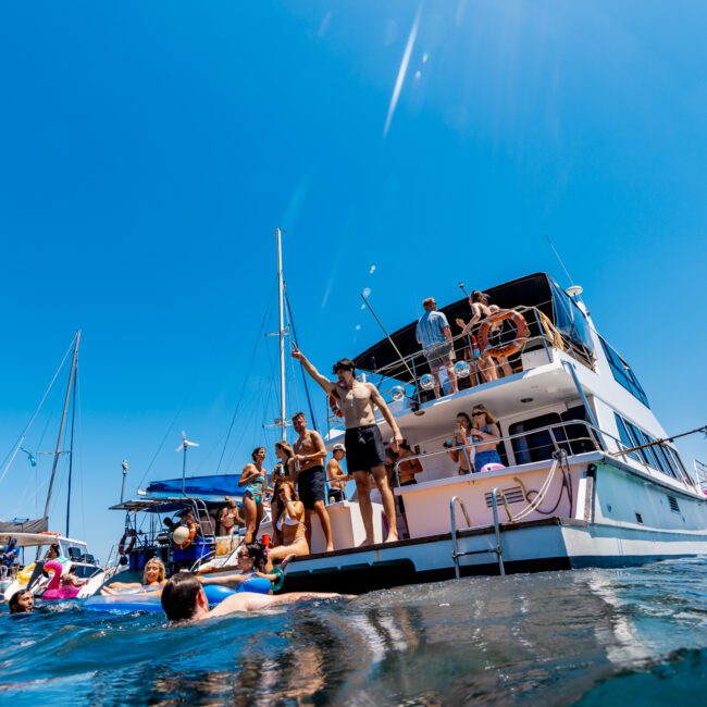 People gathered on and around a yacht enjoying the water on a sunny day. Some are swimming, while others stand or sit on the deck. The sky is clear blue, and there are several other boats in the background.