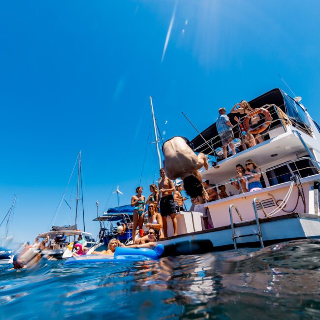 A group of people enjoy a sunny day on a yacht and in the water. One person is mid-dive off the yacht, while others relax on inflatable floats. Several boats are anchored nearby under a clear blue sky.