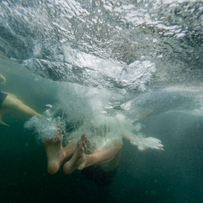 A person underwater, creating a splash with their feet as they descend into the water. The surface above is disturbed with bubbles and ripples, indicating recent movement. The water is a deep green, suggesting a natural body of water.