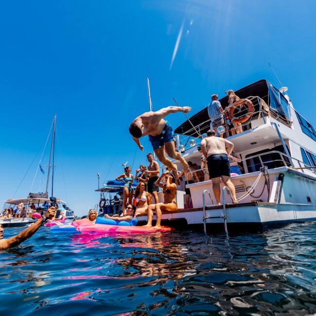 People are enjoying a sunny day on and around a yacht. Some are swimming in the water, while others are on the boat. A man is mid-air, jumping into the sea. The sky is clear and blue, creating a lively and fun atmosphere.