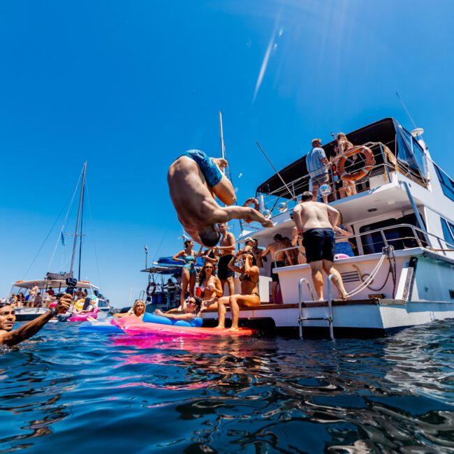 A group of people enjoy a sunny day on a boat. One person is captured mid-air while flipping into the water surrounded by others swimming and relaxing on inflatables. Several people are on the boat, and the sky is clear blue.