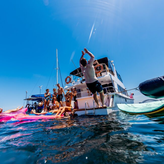 A group of people enjoying a sunny day on the water near a yacht. One person is jumping off the yacht into the ocean, while others relax on a pink inflatable and paddleboard. The sky is clear and bright blue.