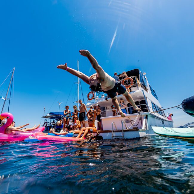 A person dives off a yacht into clear blue water. Nearby, people relax on a large inflatable pink flamingo float. Other yachts and sunbathers are in the background under a bright blue sky.
